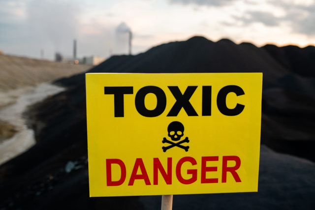 Notice on yellow board announcing toxic and dangerous area behind it with hills, polluted air and river and industrial fumes from factories