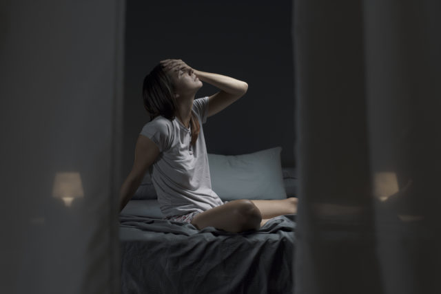 Tired woman sitting in bed at night with open window, she is suffering from the heat and she is unable to sleep