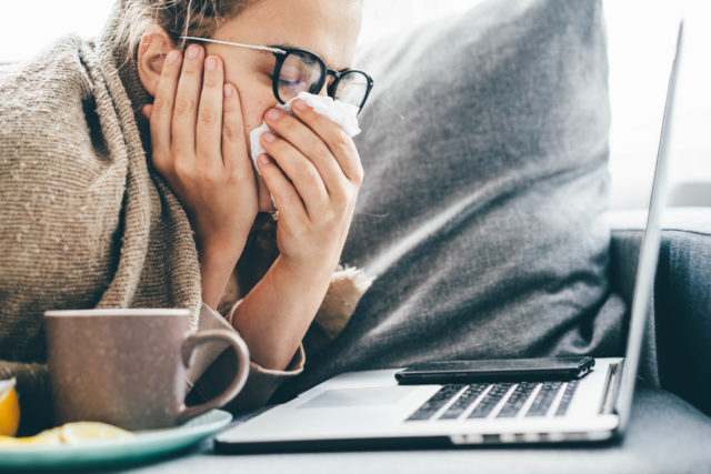 Woman blowing nose, holding handkerchief, sneezing during working with laptop in living room at home.