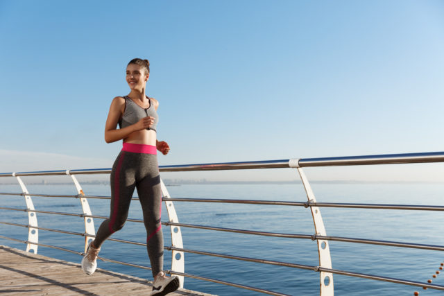 Outdoor shot of happy fitness woman running along seaside and smiling.