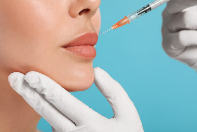 Doctor giving lips injection to young woman on light blue background, closeup. Cosmetic surgery